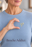 Broche cœur dorée portée sur un pull bleu clair avec un geste de la main en forme de cœur